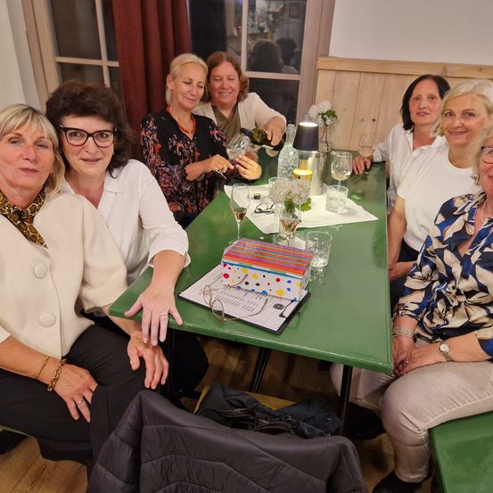 Eine Gruppe von Frauen sitzt um einen grünen Tisch, einige mit Brille, lächeln. Eine Frau gießt Wein ein, und eine Lampe, Gläser und eine bunte Schachtel stehen auf dem Tisch. Eine andere Frau hält eine Weinflasche.