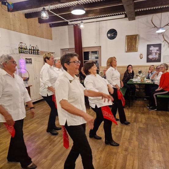 Eine Gruppe von Frauen in weißen Hemden und schwarzen Hosen tanzt in einem rustikalen Raum mit Holzböden. Dahinter sind Tische mit sitzenden Personen. An der Wand hängen Bilderrahmen und Hirschgeweihe.