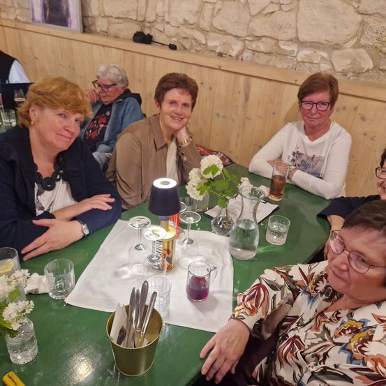 Eine Gruppe von Frauen sitzt um einen grünen Tisch herum, lächelt und posiert für ein Foto. Auf dem Tisch sind Blumen, Gläser und eine kleine Lampe. Dahinter befindet sich eine Steinmauer.