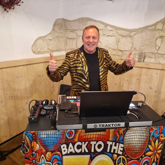 Ein Mann steht hinter einem DJ-Pult mit Laptop und Soundequipment, trägt einen goldenen Paillettenmantel und gibt ein Daumenhoch.