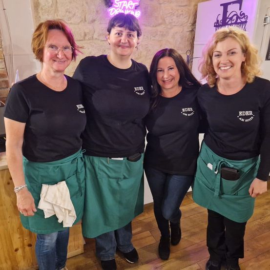 Vier Frauen in schwarzen T-Shirts und grünen Schürzen lächeln für ein Foto in einer Küche. Ein Neonschild sagt Start Drinking.