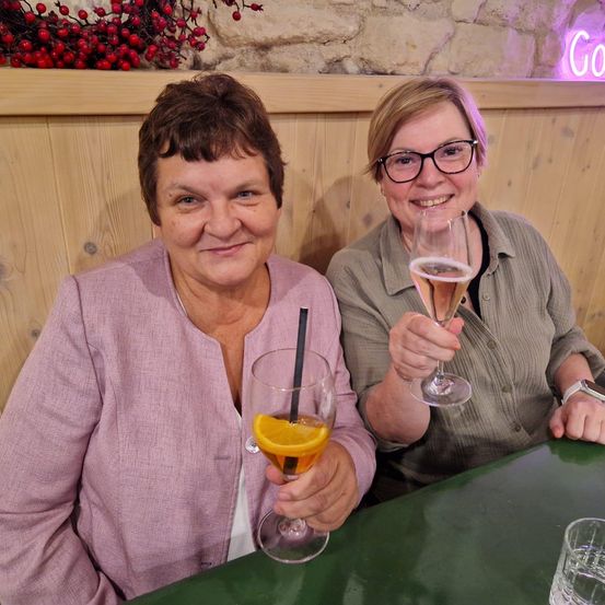 Zwei Frauen sitzen an einem Tisch und lächeln. Eine hält ein Weinglas mit Orangensaft, die andere ein Champagnerglas. Ein Neonschild mit der Aufschrift 'Go' befindet sich hinter ihnen.