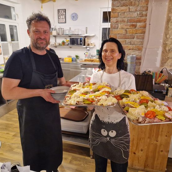 Ein Mann und eine Frau stehen in einer Küche. Der Mann trägt eine Schürze und hält einen Tablett mit Essen. Die Frau trägt eine Katzenschürze und hält einen weiteren Tablett mit Essen. Im Hintergrund befindet sich eine Ziegelwand und eine Mikrowelle.