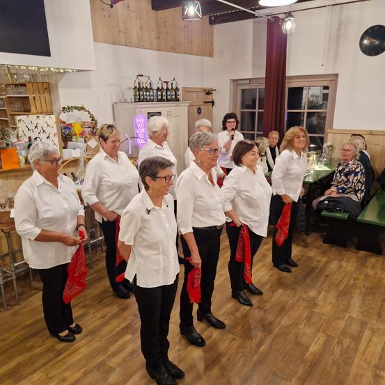 Eine Gruppe von Frauen, die weiße Hemden und schwarze Hosen tragen, steht in einem Raum mit Holzböden. Jede von ihnen hält ein rotes Tuch. Hinter ihnen befinden sich Tische mit grünen Bänken und eine Frau steht in der Nähe eines Fensters.