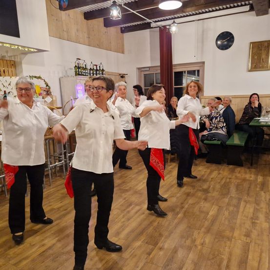 Eine Gruppe von Frauen in weißen Hemden und schwarzen Hosen tanzt in einem Restaurant. Sie tragen rote Halstücher. Der Raum hat Holzböden.
