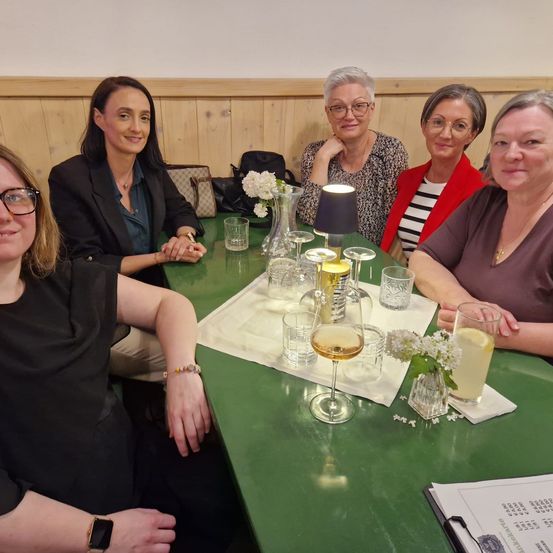 Fünf Frauen sitzen lächelnd um einen grünen Tisch herum, bereit für ein Foto. Auf dem Tisch stehen Gläser, eine Vase mit Blumen und eine leuchtende Lampe. Ein Menü ist auf der rechten Seite sichtbar.