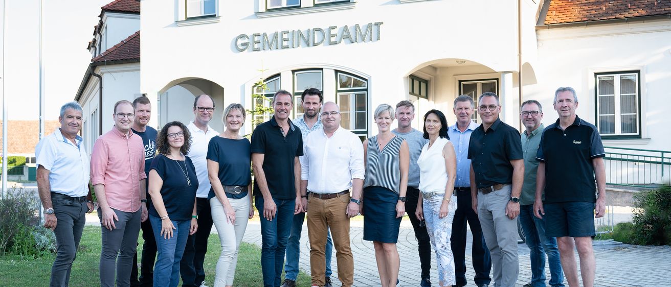 Eine Gruppe von Menschen posiert für ein Foto vor einem Gebäude mit dem Wort 'GemeinDeAMT' darauf. Sie tragen alle Freizeitkleidung.