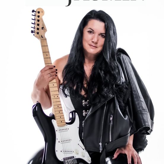 Eine Frau mit langen schwarzen Haaren, die eine schwarze Lederjacke trägt, hält eine schwarze und weiße E-Gitarre und posiert für ein Foto vor weißem Hintergrund.