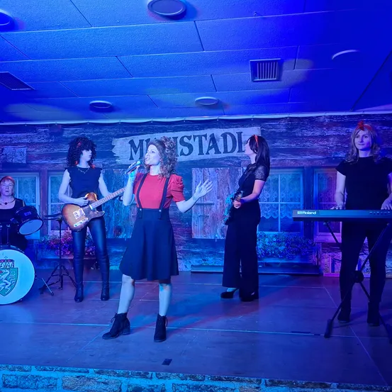 Eine Band von Frauen tritt auf der Bühne auf, mit einer Sängerin vorne, einer Gitarristin links und zwei Keyboarderinnen rechts. Die Bühne ist mit blauem Licht beleuchtet.