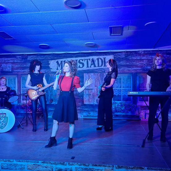 Eine Band von Frauen tritt auf der Bühne auf, mit einer Sängerin vorne, einer Gitarristin links und zwei Keyboarderinnen rechts. Die Bühne ist mit blauem Licht beleuchtet.