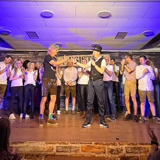 Eine Gruppe von Menschen auf der Bühne, einer hält ein Mikrofon, während andere klatschen und zusehen. Die Bühne ist mit lila Lichtern beleuchtet und hat eine Steinwand als Hintergrund.