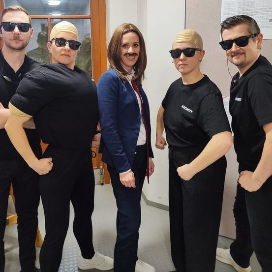 Fünf Personen, die in Schwarz gekleidet sind, stehen zusammen. Zwei haben Perücken und Schnurrbärte, und zwei haben weiße Schuhe. Eine Frau in einer blauen Blazerjacke steht in der Mitte.