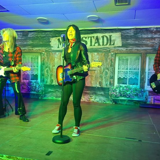 Drei Musiker spielen auf einer Bühne mit Gitarren. Eine Frau in der Mitte singt in ein Mikrofon, während zwei andere hinter ihr spielen. Im Hintergrund befindet sich ein hölzernes Schild, das 'M STADT' steht.