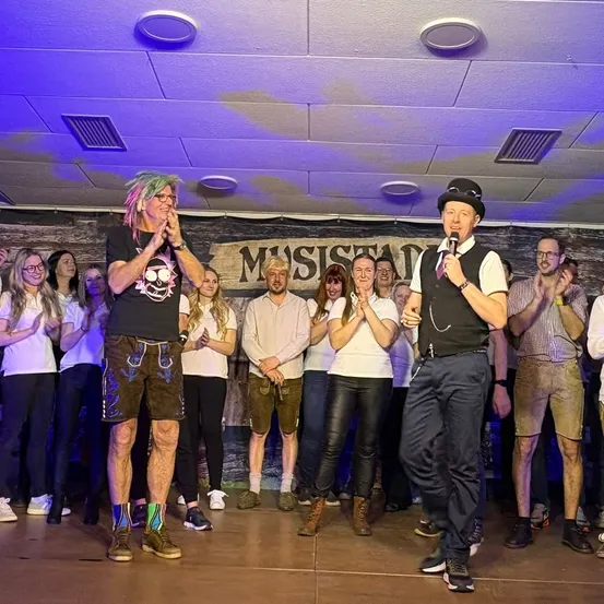 Eine Gruppe von Menschen steht auf einer Bühne unter lila Beleuchtung. Zwei Männer, einer in einem schwarzen Vest, sprechen in Mikrofone. Dahinter stehen Menschen und klatschen. Ein Banner mit der Aufschrift 'Musisian'.
