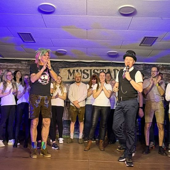 Eine Gruppe von Menschen steht auf einer Bühne unter lila Beleuchtung. Zwei Männer, einer in einem schwarzen Vest, sprechen in Mikrofone. Dahinter stehen Menschen und klatschen. Ein Banner mit der Aufschrift 'Musisian'.