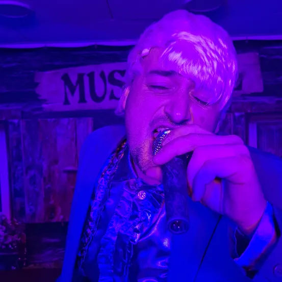 Ein Mann mit weißen Haaren singt in ein Mikrofon in einem lila beleuchteten Raum. Er trägt einen blauen Anzug und ein lila Hemd. Im Hintergrund ist ein Schild mit der Aufschrift 'Mus' zu sehen.