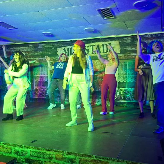 Eine Gruppe von Menschen tritt auf einer Bühne mit bunter Beleuchtung auf. Sie sind in verschiedenen Kostümen gekleidet. Die Frau vorn trägt einen roten Hut und weiße Hosen. Hinter ihr steht ein Mann in einem blauen Shirt. Es gibt eine Holzwand mit einem Schild.