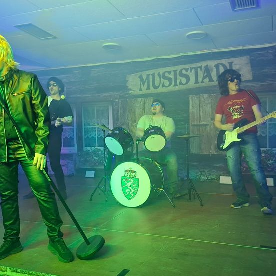 Eine Band spielt auf einer Bühne mit grüner Beleuchtung. Der Leadsänger trägt eine grüne Lederjacke. Hinter ihm spielen zwei Gitarristen. Ein Schlagzeuger sitzt vor einem Schlagzeug mit einem grünen Löwenemblem. Ein Schild steht MUSISTAD.