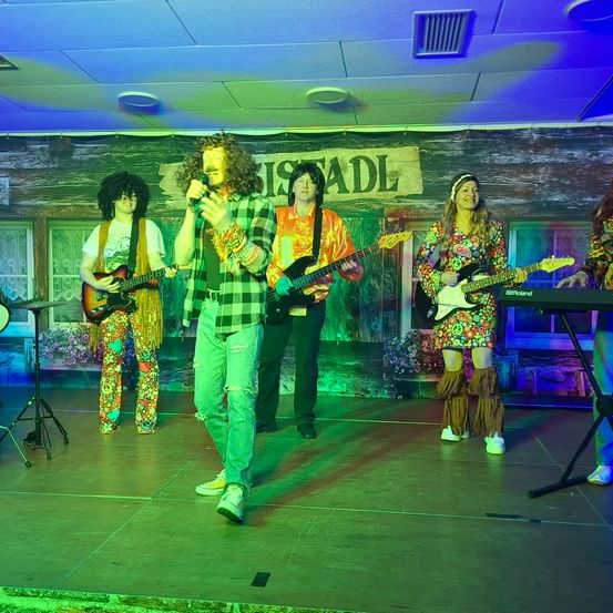 Eine Band mit Musikern in farbenfrohen, lebhaften Outfits tritt auf einer Bühne mit grüner Beleuchtung auf. Eine Person singt in ein Mikrofon, während eine andere die Gitarre spielt. Hinter ihnen befindet sich eine Kulisse mit einem Schild, das 'USTADL' steht.