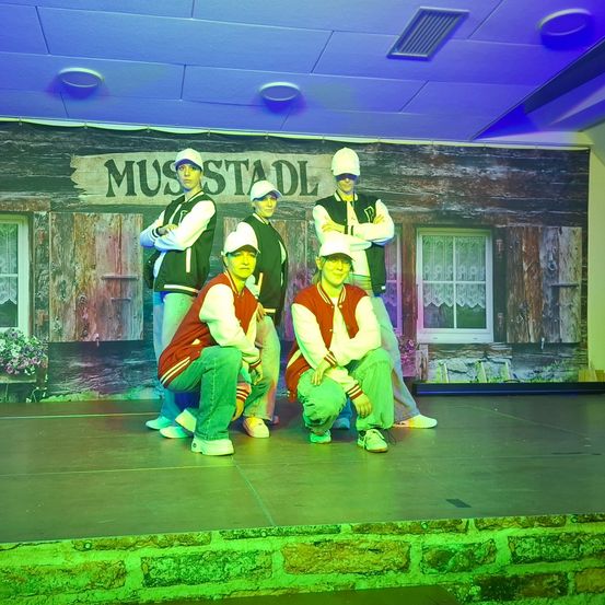 Fünf Personen in identischen Outfits posieren lächelnd auf der Bühne. Hinter ihnen befindet sich eine Kulisse mit einem hölzernen Gebäude, Fenstern und einem Schild mit der Aufschrift 'MUUSTADL'. Die Bühne ist mit grünen und violetten Lichtern beleuchtet.
