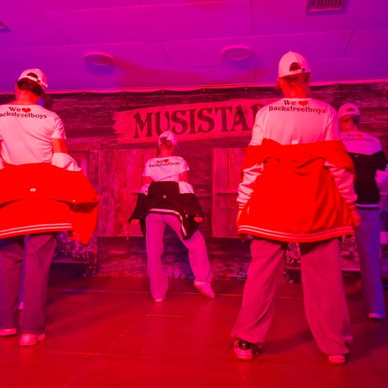 Vier Personen in roten und weißen Shirts stehen auf einer Bühne mit lila Beleuchtung. Sie tragen Mützen und Turnschuhe. Einer hält eine Jacke, ein anderer ist gebückt. Ein Schild steht 'MUSISTA'.