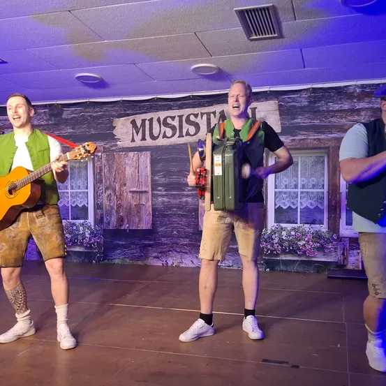 Drei Männer treten auf einer Bühne mit einer hölzernen Kulisse auf. Der Mann auf der linken Seite spielt Gitarre, der Mann in der Mitte spielt Akkordeon und der Mann auf der rechten Seite singt.
