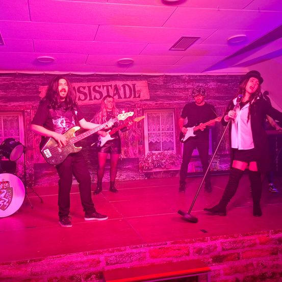 Eine Band tritt auf der Bühne mit lila Licht auf. Der Leadsänger hält ein Mikrofon, während die Gitarristen ihre Gitarren spielen. Dahinter befindet sich eine Kulisse mit einem Schild, auf dem 'SISTADL' steht.