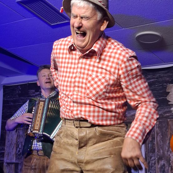Ein Mann in einem rot-weiß karierten Hemd und brauner Hose tritt auf der Bühne auf, während ein Akkordeonspieler im Hintergrund spielt.