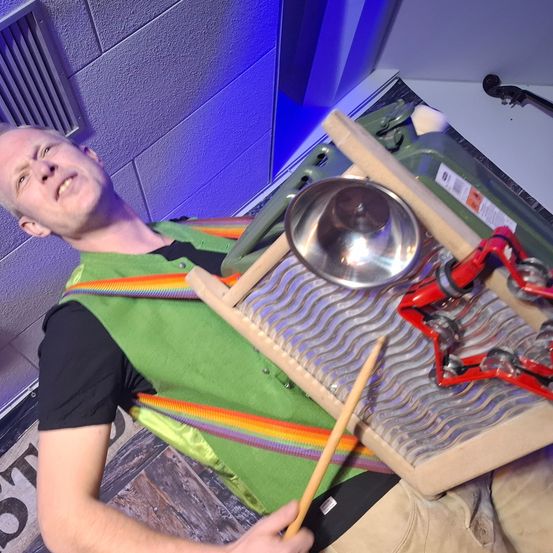 Ein Mann in einer grünen Weste und beigefarbener Hose spielt ein Musikinstrument mit einem Drumstick. Das Instrument ist ein metallisches, wellenförmiges Objekt mit einer Becken und roten Maracas.