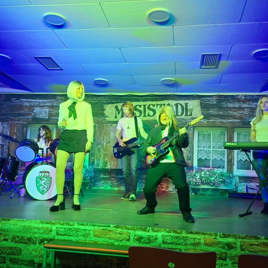 Eine Band auf der Bühne mit einer Sängerin, einem Gitarristen, einem Bassisten und einer Schlagzeugerin. Die Bühne hat einen rustikalen Holzhintergrund.