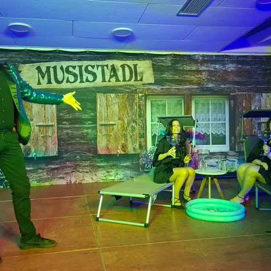 Eine Bühnenaufstellung mit einem hölzernen Hintergrund, beschriftet mit 'MUSISTADL'. Ein Mann in Grün zeigt auf zwei Frauen, eine hält ein Weinglas und sitzt auf einer Liege, die andere sitzt in der Nähe.