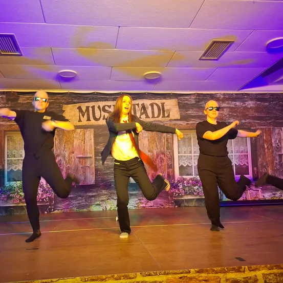 Drei Personen in schwarzen Anzügen und Sonnenbrillen führen eine Tanzroutine auf der Bühne auf, mit einem Hintergrund eines hölzernen Schilds, das 'MUSTALD' steht.