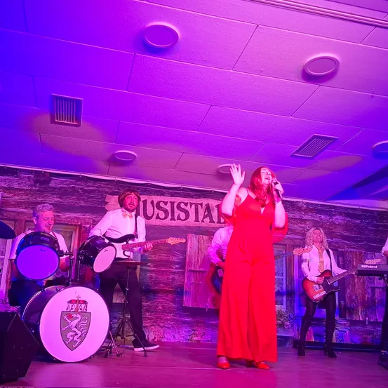 Eine Band tritt auf der Bühne unter lila Lichtern auf. Eine Frau in einem roten Kleid singt in ein Mikrofon. Hinter ihr spielt ein Gitarrist. Auf der linken Seite spielen ein Schlagzeuger und ein Bassist ihre Instrumente.