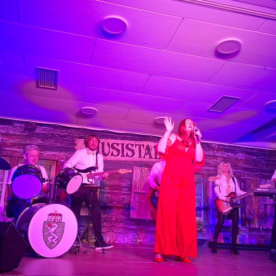 Eine Band tritt auf der Bühne unter lila Lichtern auf. Eine Frau in einem roten Kleid singt in ein Mikrofon. Hinter ihr spielt ein Gitarrist. Auf der linken Seite spielen ein Schlagzeuger und ein Bassist ihre Instrumente.