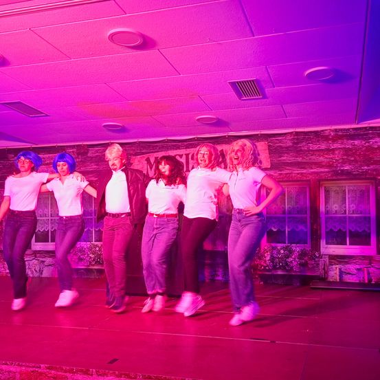 Eine Gruppe von Menschen in weißen Shirts und Jeans tritt auf einer Bühne mit lila Beleuchtung auf, einer trägt eine blaue Perücke.