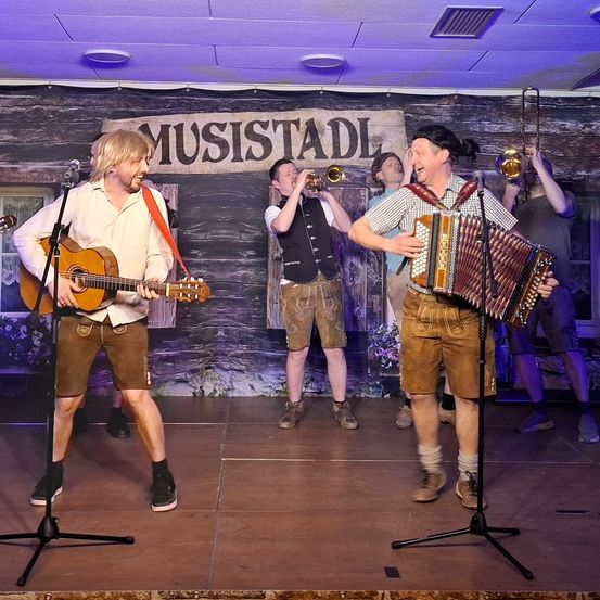 Fünf Musiker treten auf einer Holzbühne mit einem Schild auf, das 'MUSISTADL' steht. Einer spielt Gitarre, ein anderer Akkordeon und zwei weitere spielen Blechblasinstrumente.