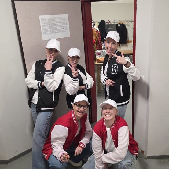 Fünf Frauen in passenden Jacken posieren für ein Foto in einem Raum mit einem Spiegel und einer offenen Tür. Sie tragen alle weiße Kappen und lächeln.