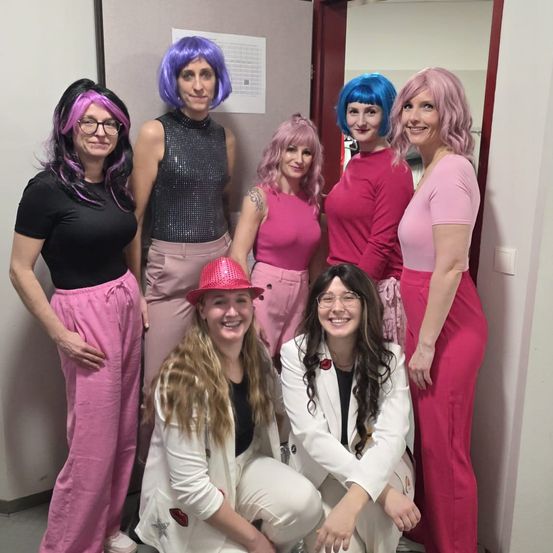 Eine Gruppe von Frauen in rosa Outfits posiert für ein Foto. Einige tragen Perücken und Hüte. Sie lächeln und stehen oder sitzen in einem Raum.