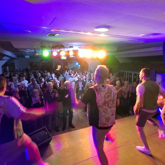 Eine Gruppe älterer Erwachsener tanzt auf einer Bühne unter bunten Lichtern, während ein großes Publikum zuschaut.