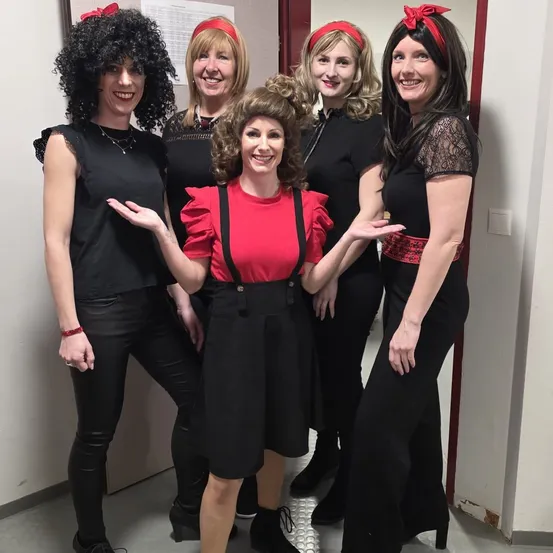 Fünf Frauen in schwarzen und roten Outfits mit roten Stirnbändern posieren für ein Foto. Eine Frau trägt ein rotes Shirt und schwarze Hose. Hinter ihnen ist eine weiße Wand mit einer roten Tür.