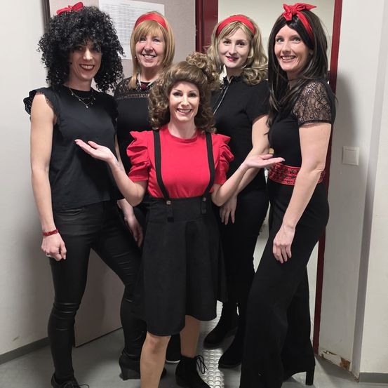 Fünf Frauen in schwarzen und roten Outfits mit roten Stirnbändern posieren für ein Foto. Eine Frau trägt ein rotes Shirt und schwarze Hose. Hinter ihnen ist eine weiße Wand mit einer roten Tür.