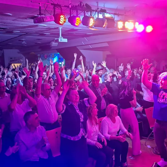 Eine Gruppe von Menschen ist in einem Raum und hebt die Hände, vielleicht jubeln sie für jemanden. Einige tragen Brillen und Hüte. Der Raum ist mit pinkfarbenen Lichtern beleuchtet und eine Projektorlampe hängt von der Decke.
