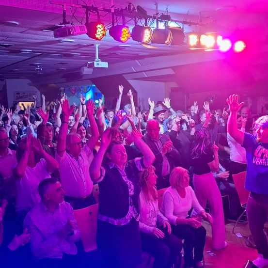 Eine Gruppe von Menschen ist in einem Raum und hebt die Hände, vielleicht jubeln sie für jemanden. Einige tragen Brillen und Hüte. Der Raum ist mit pinkfarbenen Lichtern beleuchtet und eine Projektorlampe hängt von der Decke.