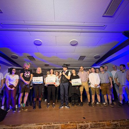 Eine Gruppe von Menschen steht mit blauen Lichtern auf der Bühne und hält Schilder mit Geldwerten. Der Mann in der Mitte spricht in ein Mikrofon.