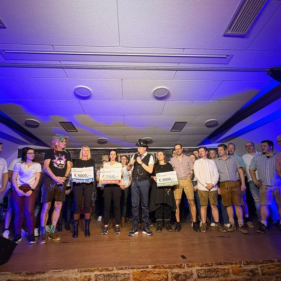 Eine Gruppe von Menschen steht mit blauen Lichtern auf der Bühne und hält Schilder mit Geldwerten. Der Mann in der Mitte spricht in ein Mikrofon.