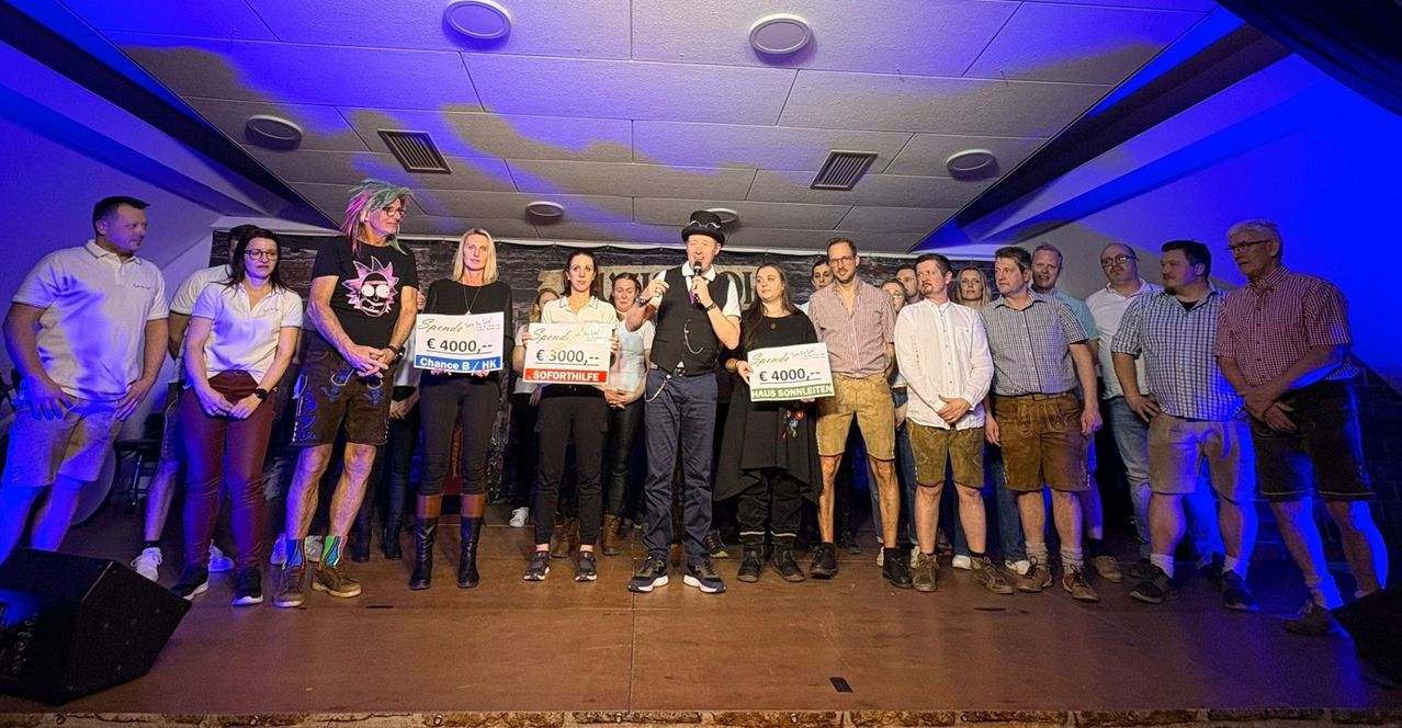 A group of people on stage holding signs with prizes. A man speaks into a microphone. The audience listens attentively. Various prizes include Spende Chance B for 4000 euros, Soforthilfe for 3000 euros, and Haus Sonnenleiten for 4000 euros.