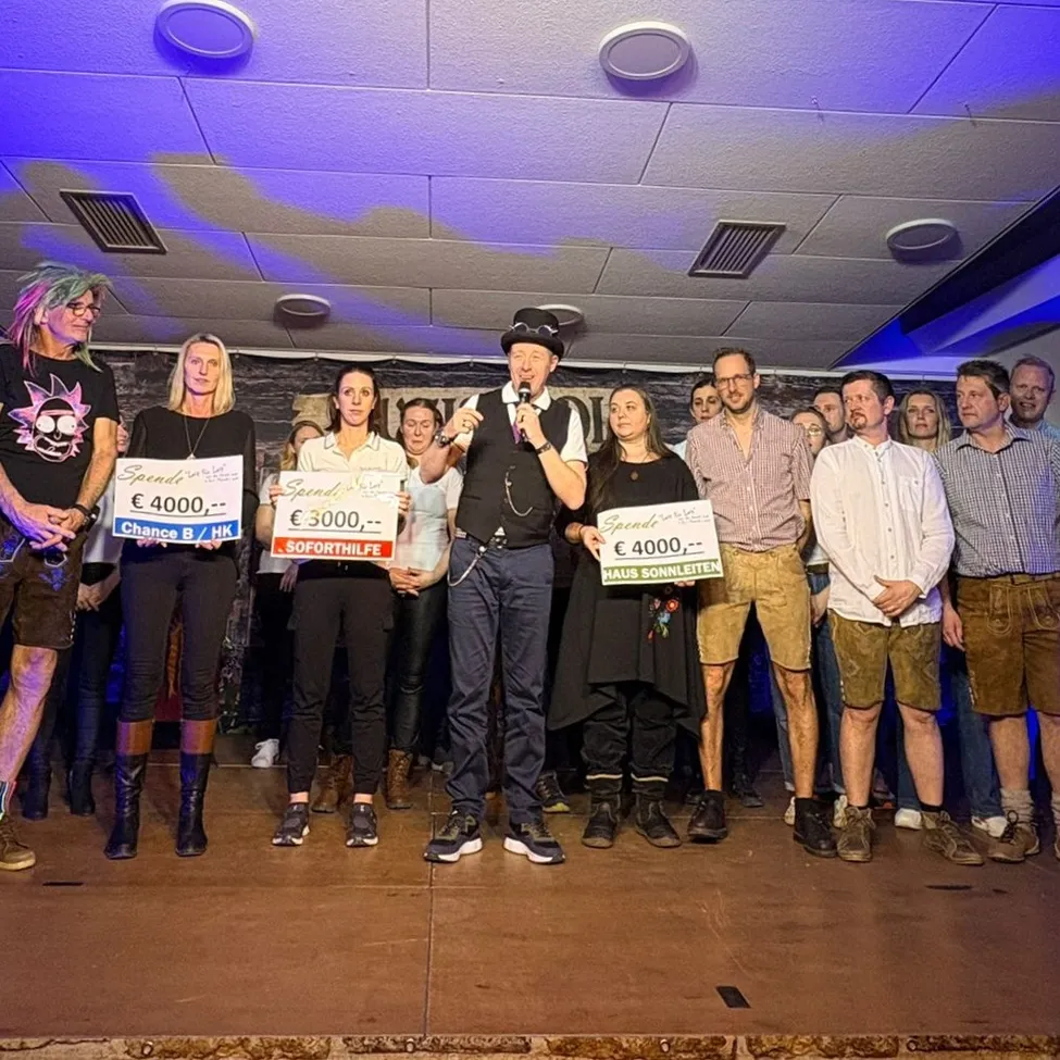 Eine Gruppe von Menschen auf der Bühne hält Schilder mit Preisen in der Hand. Ein Mann spricht in ein Mikrofon. Das Publikum hört aufmerksam zu. Verschiedene Preise umfassen Spende Chance B für 4000 Euro, Soforthilfe für 3000 Euro und Haus Sonnenleiten für 4000 Euro.