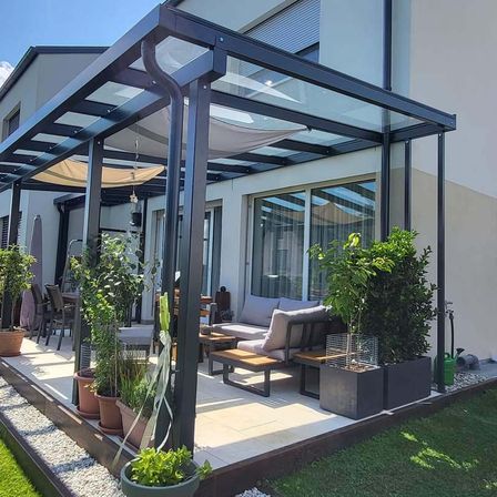Ein moderner Terrassenbereich mit Glasdach, umgeben von Pflanzen und einer Couch, mit einem Essplatz und einem Haus im Hintergrund.