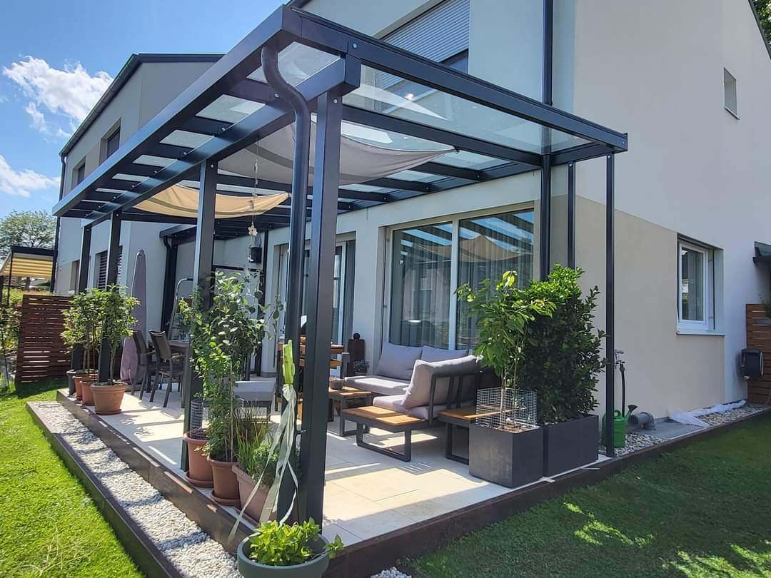 Ein moderner Terrassenbereich mit Glasdach, umgeben von Pflanzen und einer Couch, mit einem Essplatz und einem Haus im Hintergrund.