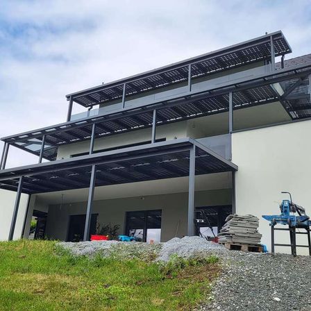 Ein modernes Haus mit Solarpanel auf dem Dach und einem Balkon davor. Ein Grasbereich mit Kies und einer blauen Maschine.
